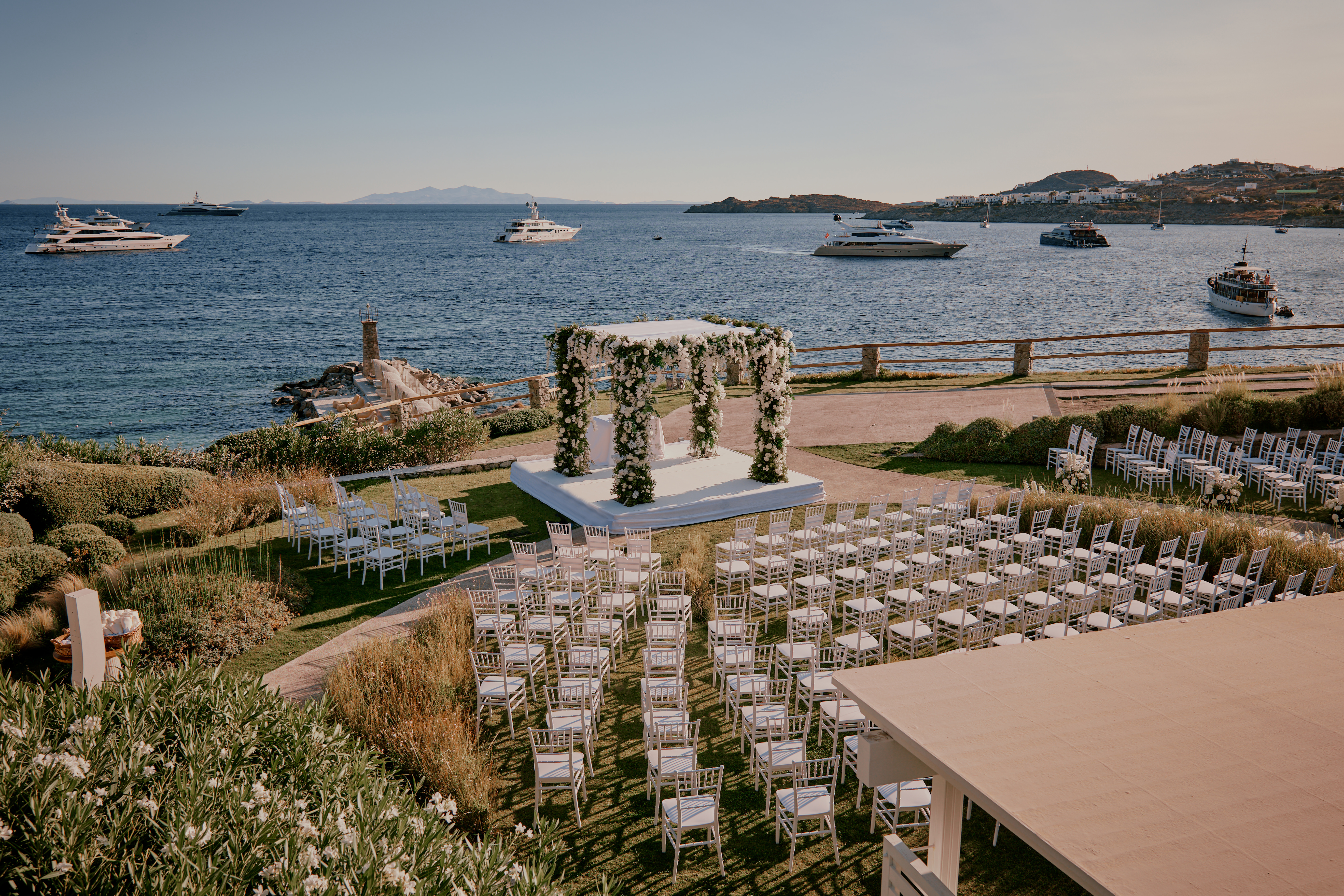 Elegant luxury wedding ceremony setup in Mykonos with white chairs arranged around a floral chuppah overlooking the Aegean Sea and anchored yachts in the background